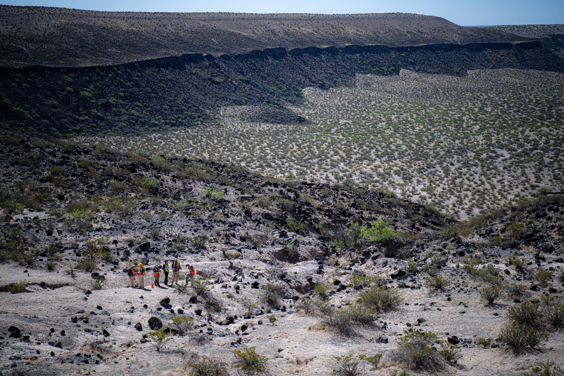 A small team of RISE 2 field researchers gathers on the pale, rocky floor of Kilbourne Hole, dwarfed by the scrub-dotted walls of the nearly two-mile-wide volcanic crater in the Potrillo Volcanic Field, New Mexico.